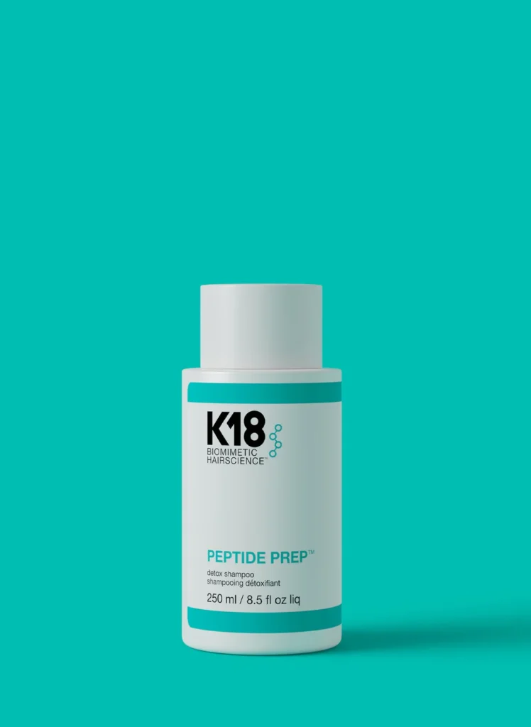 A bottle of K18 Peptide Prep Detox Shampoo on a turquoise countertop and background.