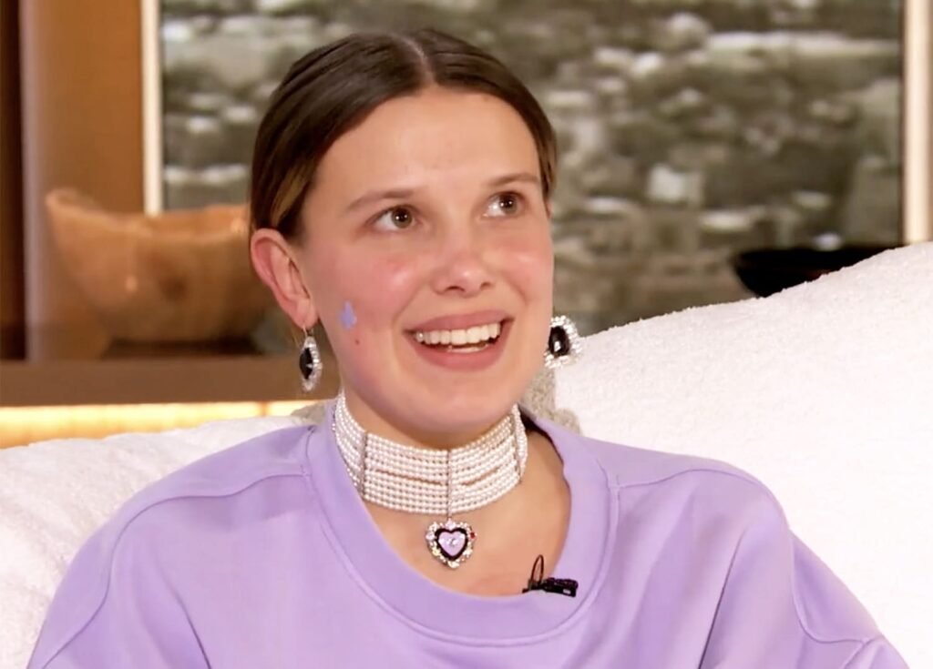 Millie Bobby Brown smiles at an event with a visible pimple patch on her cheek.